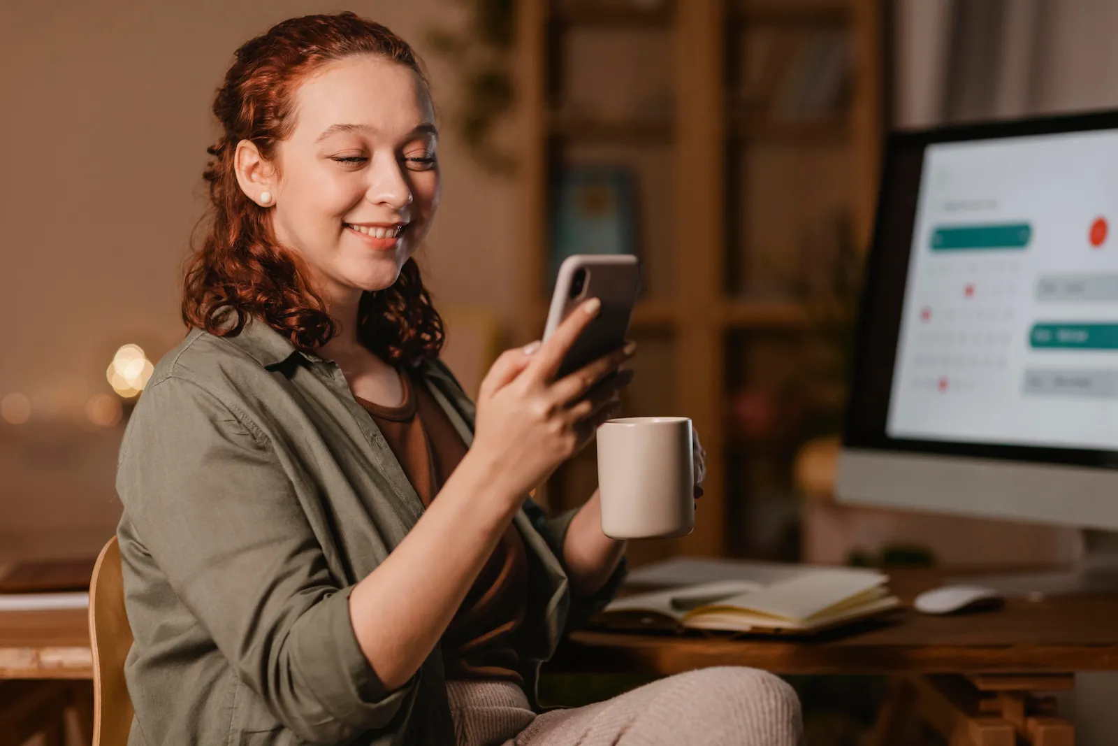 Uma mulher ruiva sorrindo, segurando uma caneca e olhando para o celular em frente a um monitor com calendário